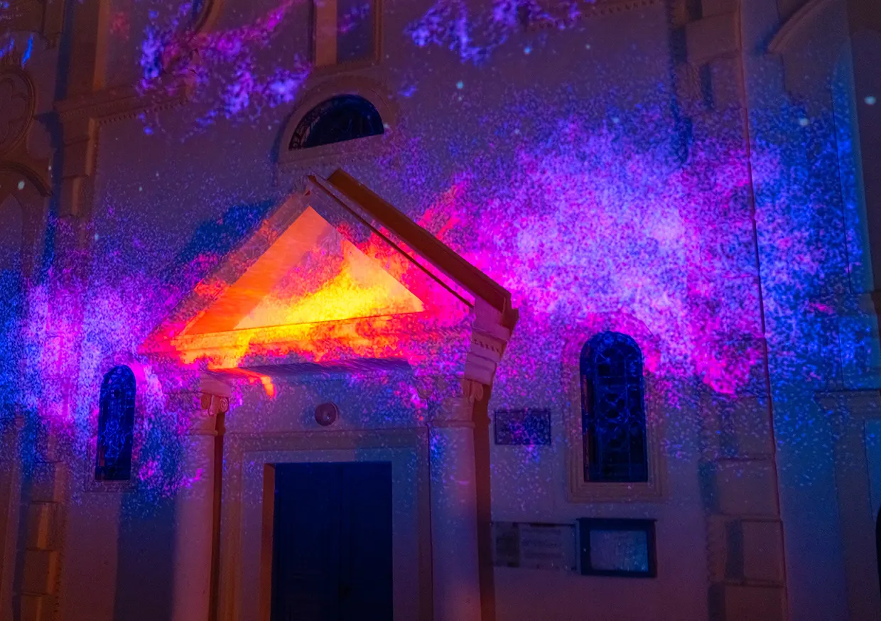 Violet particles and an orange glowing pediment projected on a church entrance at night