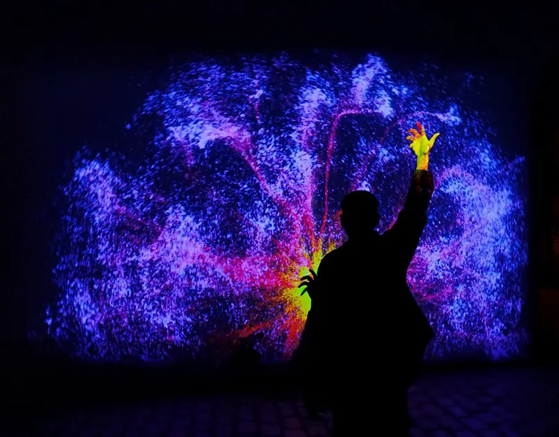 Silhouette of a visitor with a raised arm in front of a wall of glowing blue and violet particles